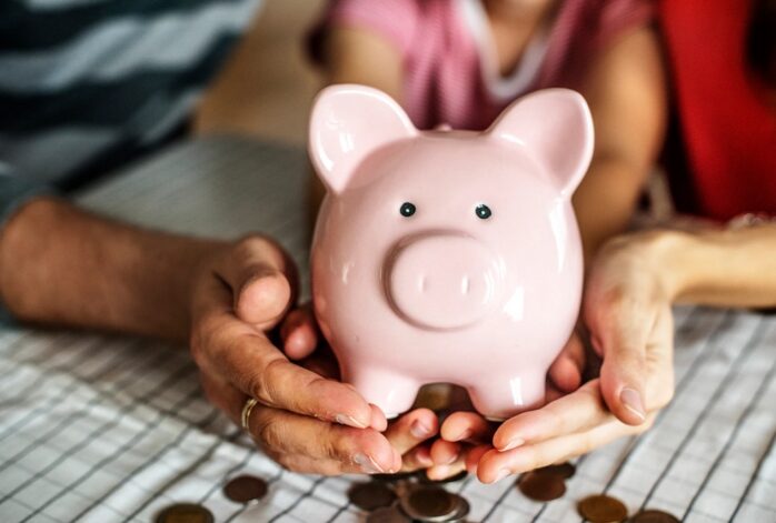 A close-up of a couple and their child’s hands holding a pink piggy bank
